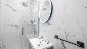Un baño moderno con pared de mármol, cabina de ducha con mampara de vidrio, lavamanos blanco con espejo y barra de apoyo.