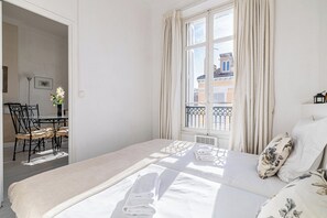 Habitación de hotel con cama, ventana con cortinas, mesa y sillas, y vista a edificios.