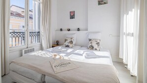 Ein Hotelzimmer mit Bett, einem Fenster mit Vorhängen, einer kleinen Ablagefläche mit Dekoration und einer Wand mit einem gerahmten Bild.
