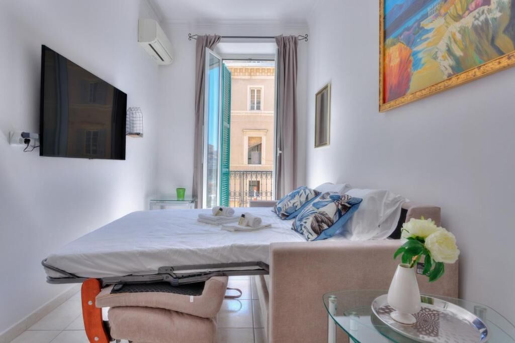 Camera d'albergo con un letto, un tavolino con un vaso di fiori, un quadro appeso al muro e vista sui palazzi attraverso una finestra aperta.