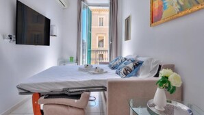 A hotel room with a bed, a glass table with a vase of flowers, a painting on the wall, and a view of buildings through an open window.