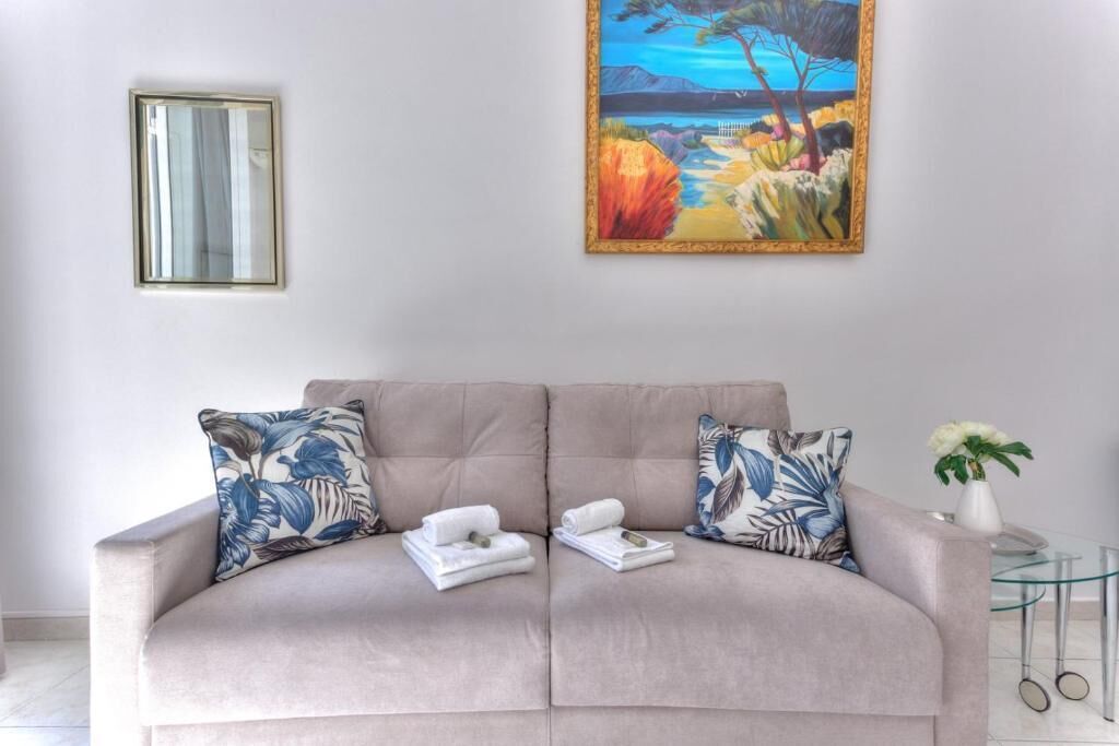 A beige sofa with floral cushions, a painting of a landscape, and a small table with a vase.