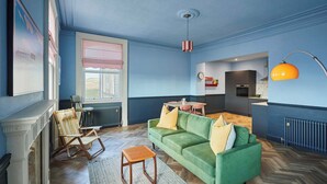 Ein Wohnzimmer mit einem grünen Sofa, einem Holztisch und einem Kamin. Ein Fenster mit gestreiftem Vorhang bietet einen Blick ins Freie.