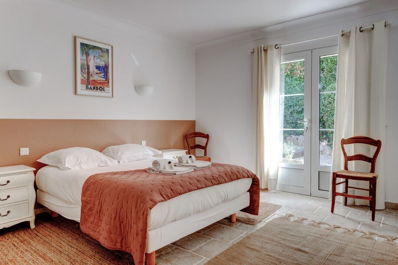 A bedroom with a bed, a nightstand, a chair, a window with curtains, and a wall-mounted light fixture.