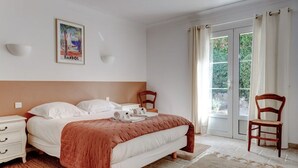 Et soveværelse med seng, natbord, stol, et vindue med gardiner og en vægmonteret lampe.