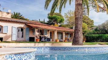 A villa with a tiled swimming pool, outdoor furniture, and palm trees.