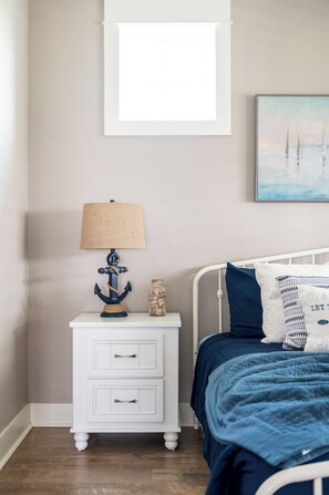 Un dormitorio con cama, una mesita de noche con lámpara, una pequeña mesa con un tarro y un cuadro en la pared.