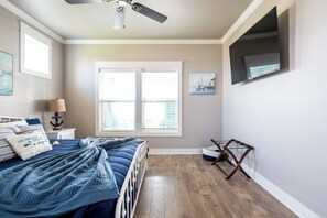 Ein Schlafzimmer mit Bett, Deckenventilator, Fenster mit Jalousien, an der Wand montiertem Fernseher und einem kleinen Tisch mit Lampe.