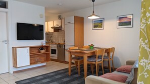 A compact living space with a kitchenette, a dining area with a table and chairs, a sofa, a TV mounted on the wall, and two framed pictures on the wall.