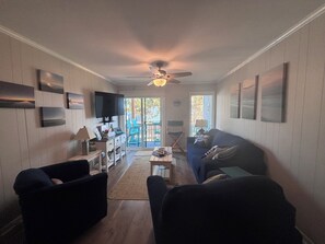 A living room with a ceiling fan, a TV, a sofa, and a coffee table. - 'Just Chillaxin'-2-bedroom condo in lovely Ocean Isle Beach, NC-oceanview & pool (Ocean Isle Beach)