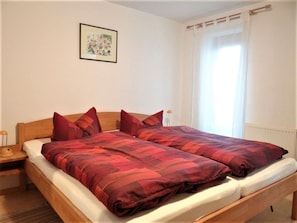 Un lit double avec une literie rouge, une table de chevet avec une lampe, un tableau encadré au mur et une fenêtre avec des rideaux.