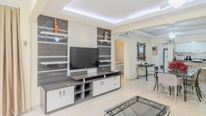 A modern living room with a flat-screen TV, built-in shelves, and a dining area with a glass table and chairs.