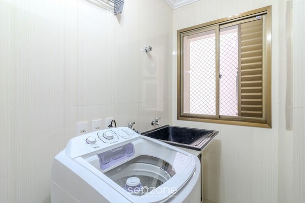A compact laundry room with a front-loading washing machine, a sink, and a window with blinds.