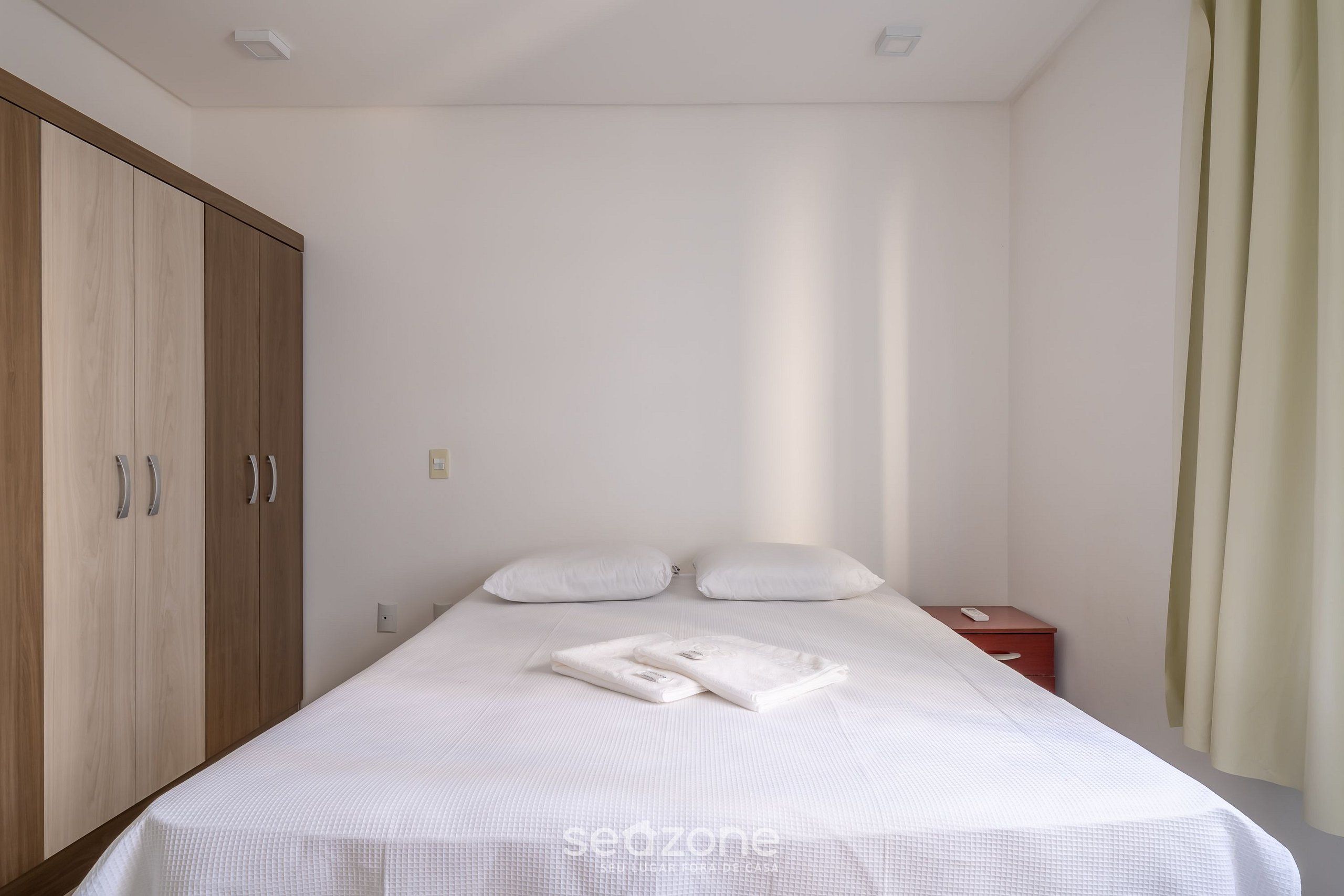 Una cama bien hecha con sábanas y almohadas blancas, un armario de madera, una mesita de noche con un teléfono y una cortina en la ventana.