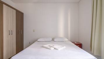 Una cama bien hecha con sábanas y almohadas blancas, un armario de madera, una mesita de noche con un teléfono y una cortina en la ventana.