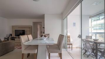 Una sala de estar moderna con una mesa de comedor de vidrio, asientos cómodos, un televisor de pantalla plana y una cocina al fondo.