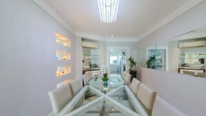 Un comedor moderno con una mesa y sillas de vidrio, una araña y una cocina visible a través de una puerta abierta.