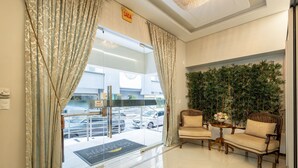 A hotel lobby with a glass door entrance, two armchairs, a small table with a vase, and a green wall with plants.