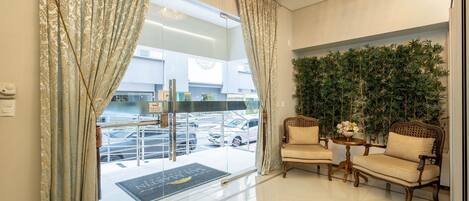 A hotel lobby with a glass door entrance, two armchairs, a small table with a vase, and a green wall with plants.