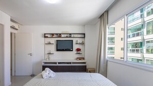 A hotel room with a bed, a TV, a desk, and a window with a view of a building.