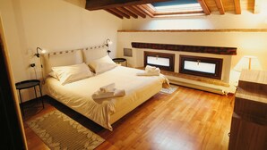 Een slaapkamer met een houten plafond, een bed met witte lakens, twee nachtkastjes, een bijzettafel, een raam met uitzicht en een ingebouwde heater.
