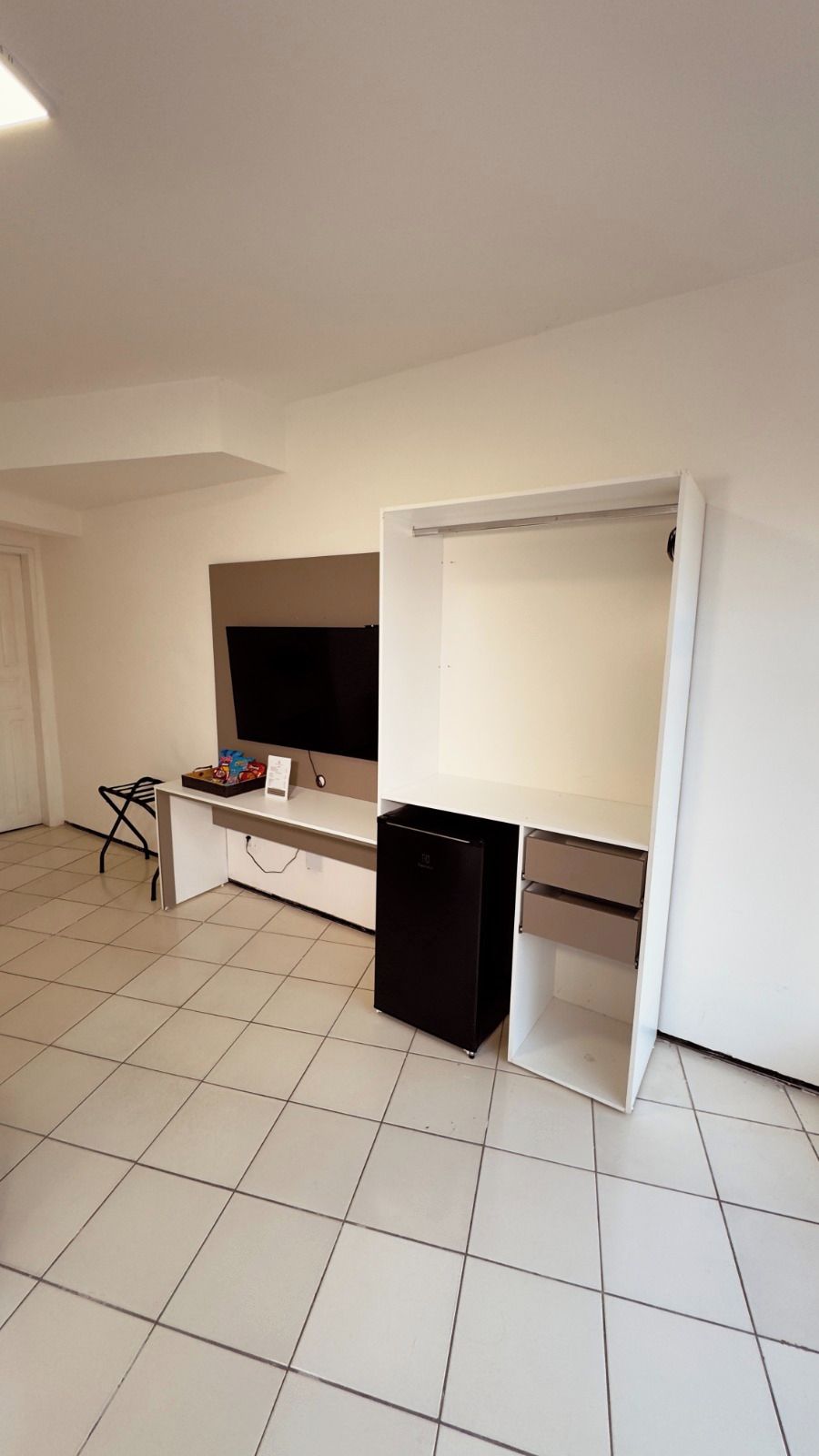 Una habitación con un escritorio blanco, un refrigerador negro y un televisor en la pared.