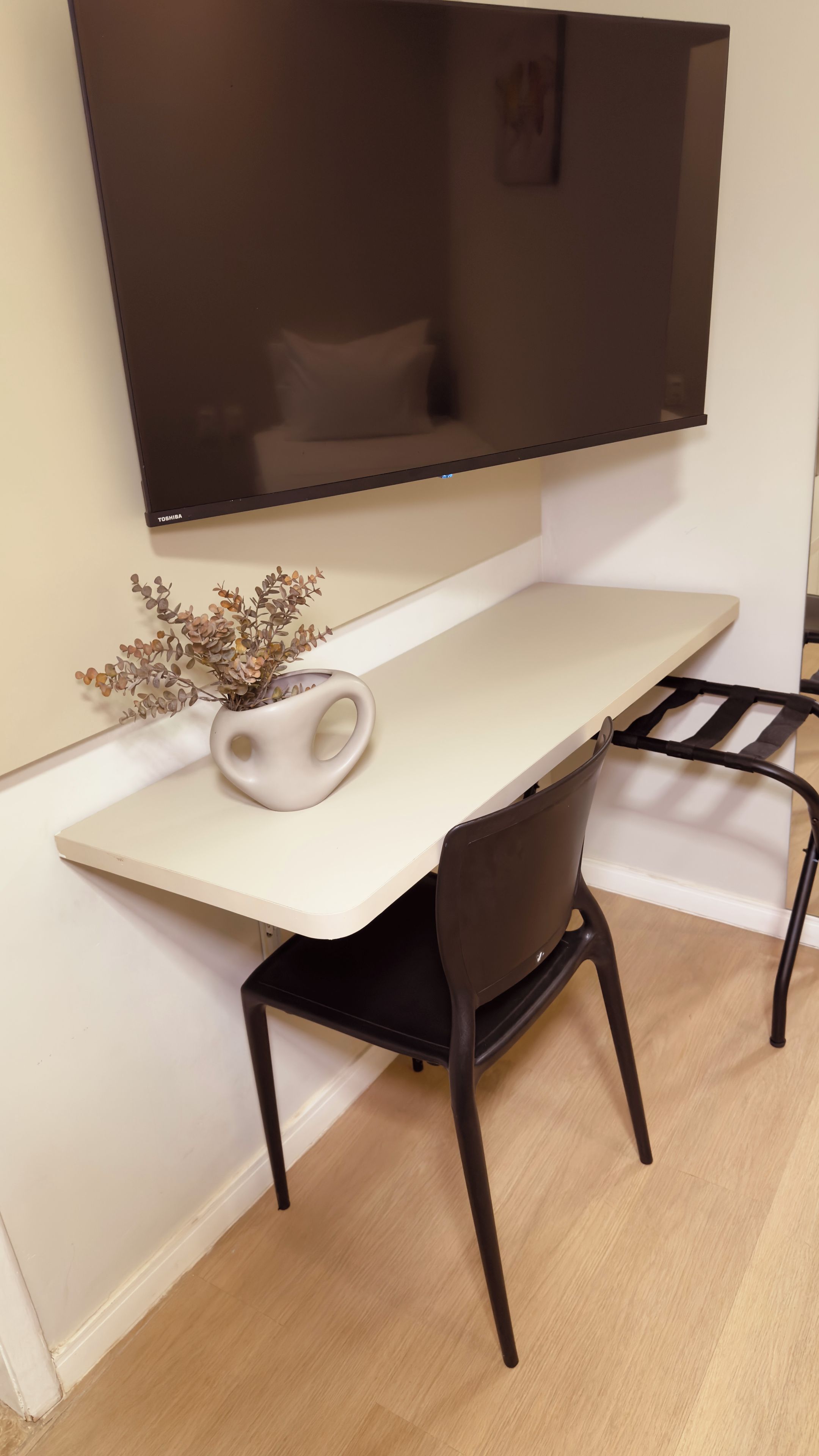 A modern desk with a mounted flat-screen TV, a vase with dried flowers, and a chair.