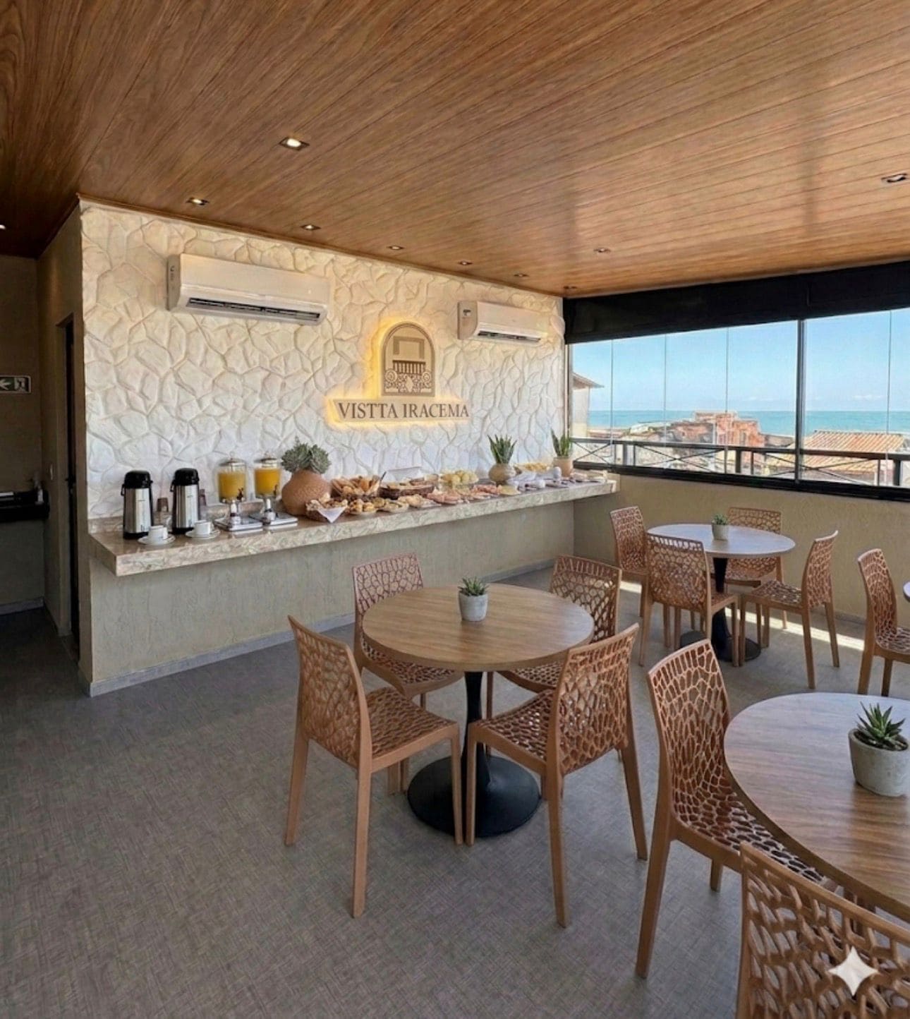 A breakfast bar with a stone wall backdrop, a view of the sea, and a seating area with round tables and chairs.