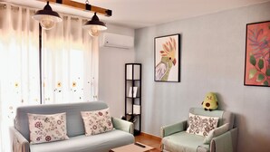 A living room with a sofa, armchair, and a small table. There is a bookshelf, a wall-mounted shelf, and a decorative pillow on the armchair.