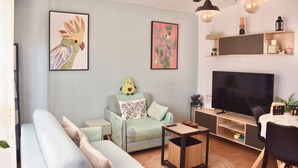 A living room with a sofa, armchair, and a television. There are wall art pieces, a lamp, and a shelf with decorative items.