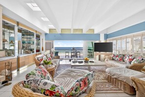 A spacious living area with wicker furniture, a large flat-screen TV, and a view of the sea.