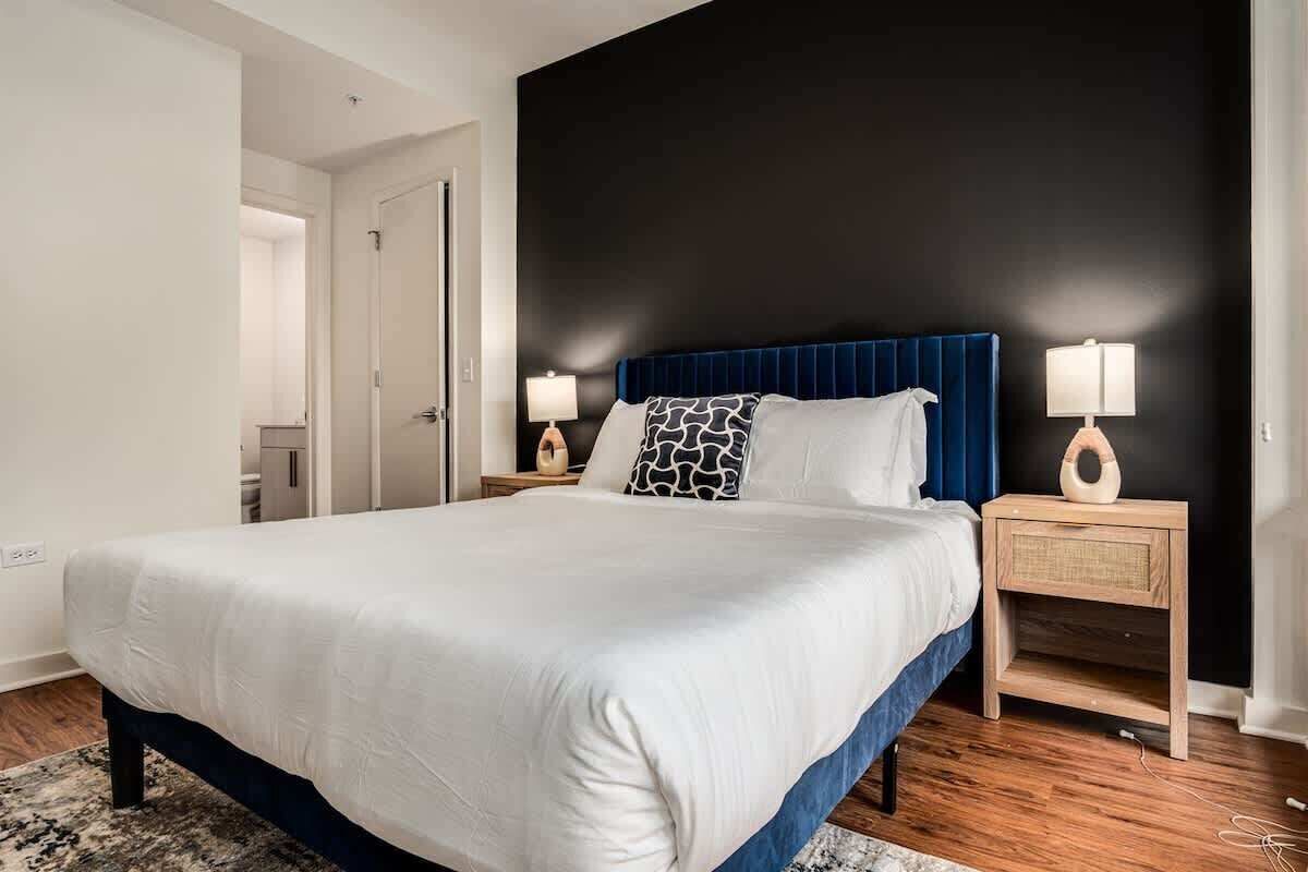 Una camera da letto moderna con un letto grande, una testiera blu scuro, biancheria da letto bianca e due comodini con lampade.