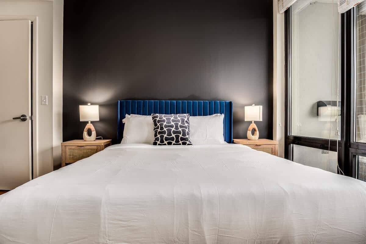 A hotel room with a large bed, two bedside tables with lamps, a dark wall, and a large window.