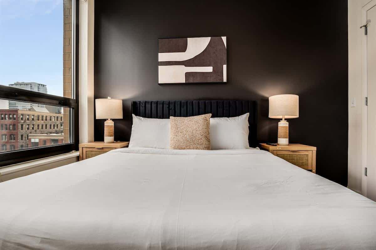 A hotel room with a large bed, two bedside lamps, a dark wall with a geometric artwork, and a window with a city view.