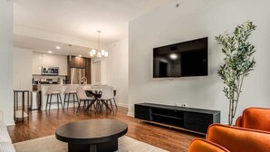 Una sala de estar moderna con una mesa de centro redonda, sillas naranjas, un televisor de pantalla plana montado en la pared y un área de comedor al fondo.