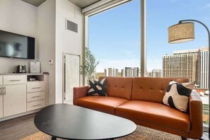 A modern living room with a leather sofa, a flat-screen TV, a coffee machine, and a large window offering a city view. - Boulevard Stays Sleek Studio Pool, Gym, & Paid Parking (Phoenix)
