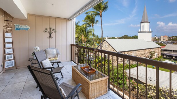 A balcony with wicker furniture, a glass-top table, and a view of a church and palm trees.