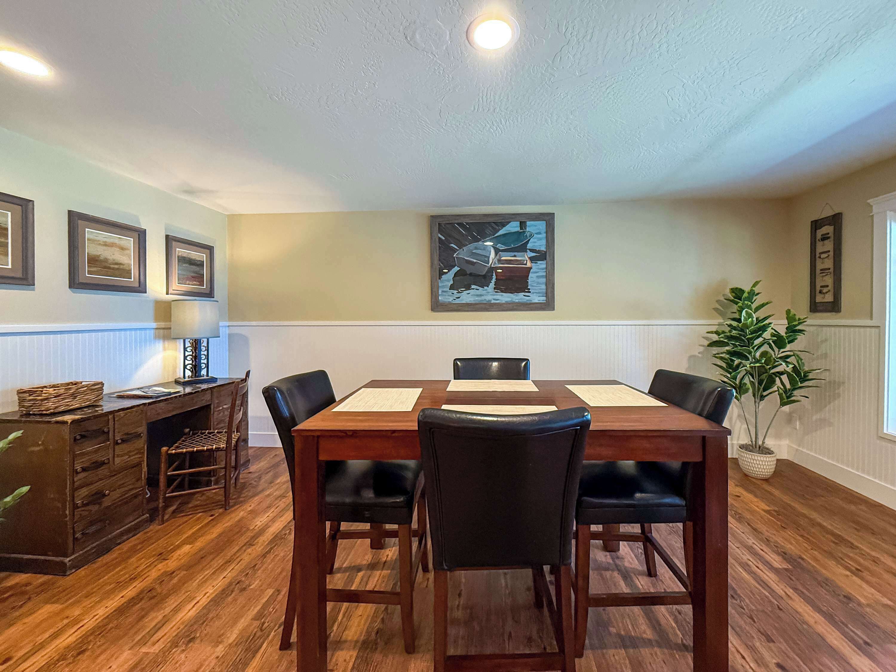 A dining area with a wooden table, four chairs, a lamp, and framed artwork on the wall.