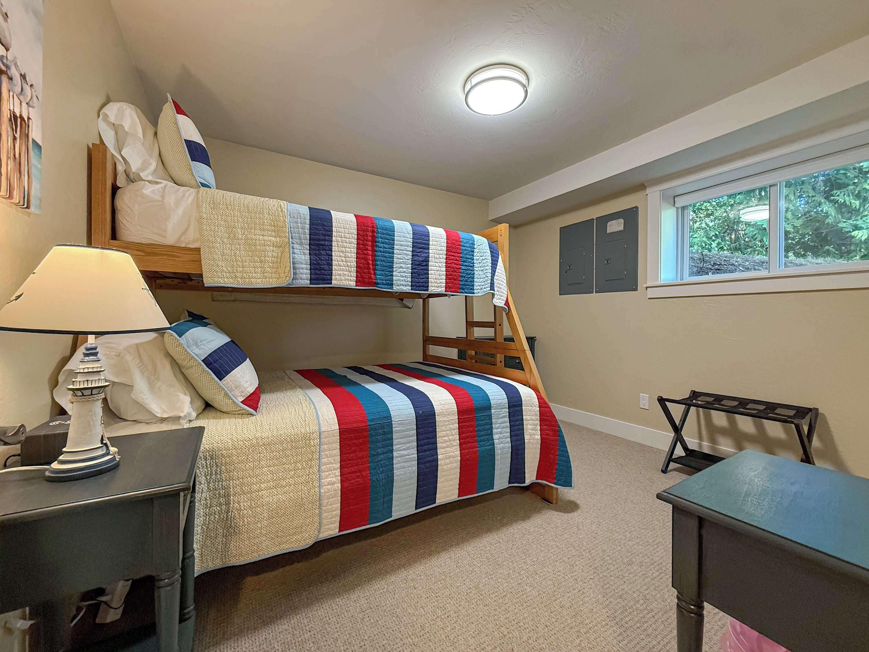 A bunk bed room with a striped bedspread, a desk, a lamp, and a window with a view of greenery.