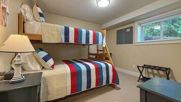 A bunk bed room with a striped bedspread, a desk, a lamp, and a window with a view of greenery.