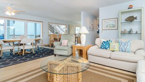 A spacious living room with a glass coffee table, a sofa with decorative pillows, a wicker chair, and a dining area with a table and chairs.