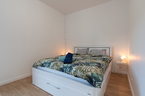 Un lit simple avec une couvre-lit à motifs de feuilles, une table de chevet blanche avec une lampe et une commode blanche.