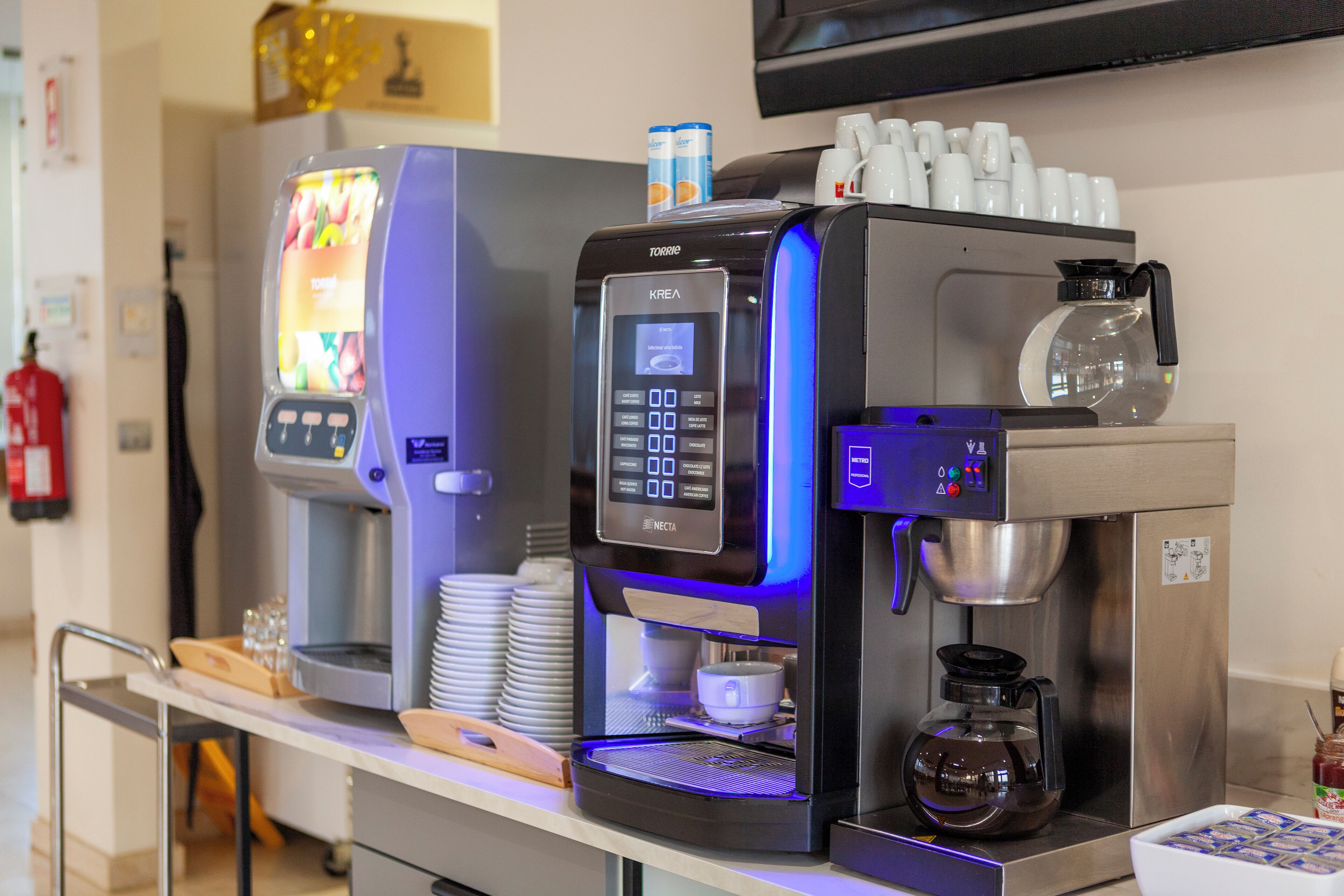 Una cafetera con panel digital, una jarra de café y una pila de vasos desechables.