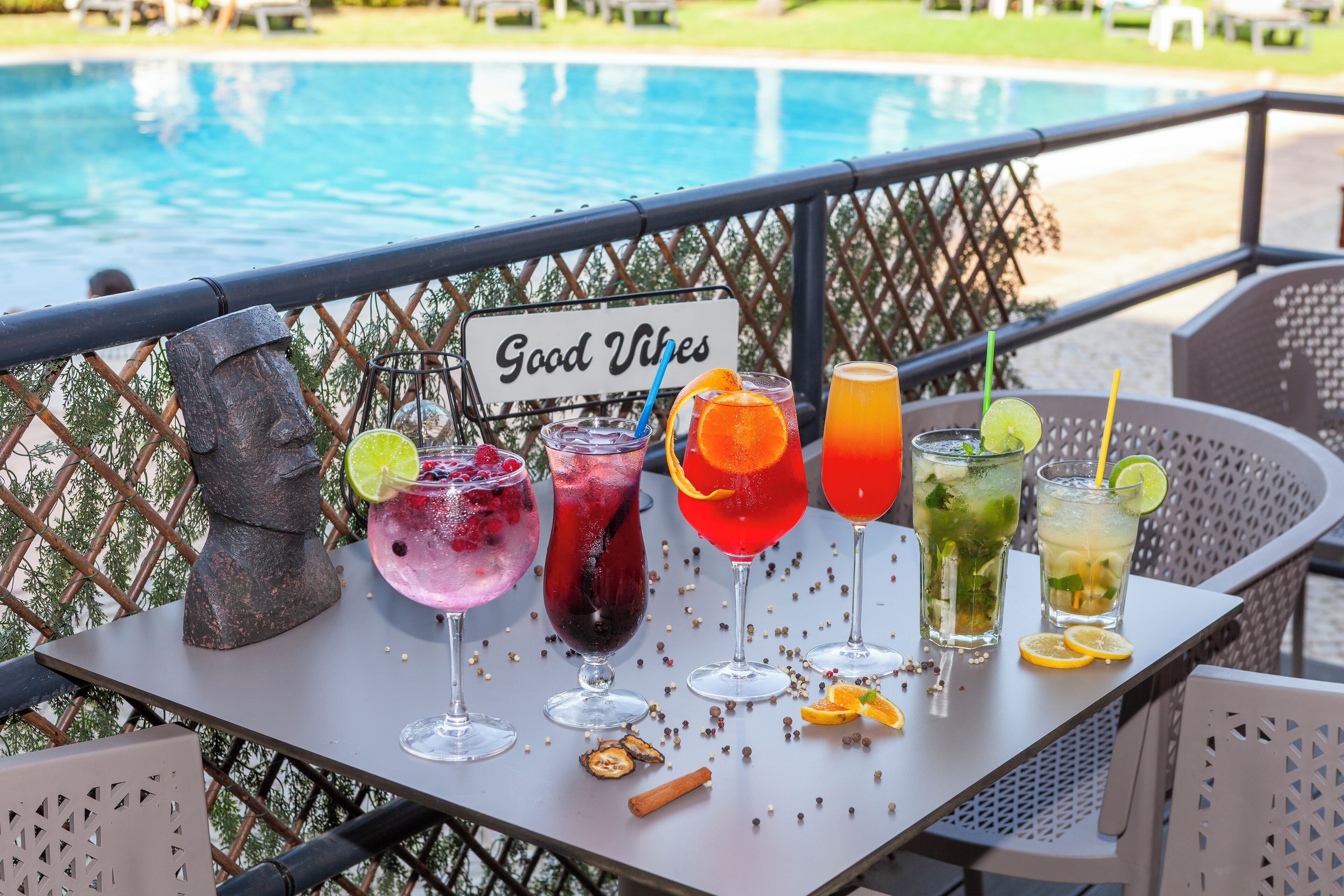 A table with various drinks and a sign that reads 'Good Vibes' on a balcony overlooking a pool.