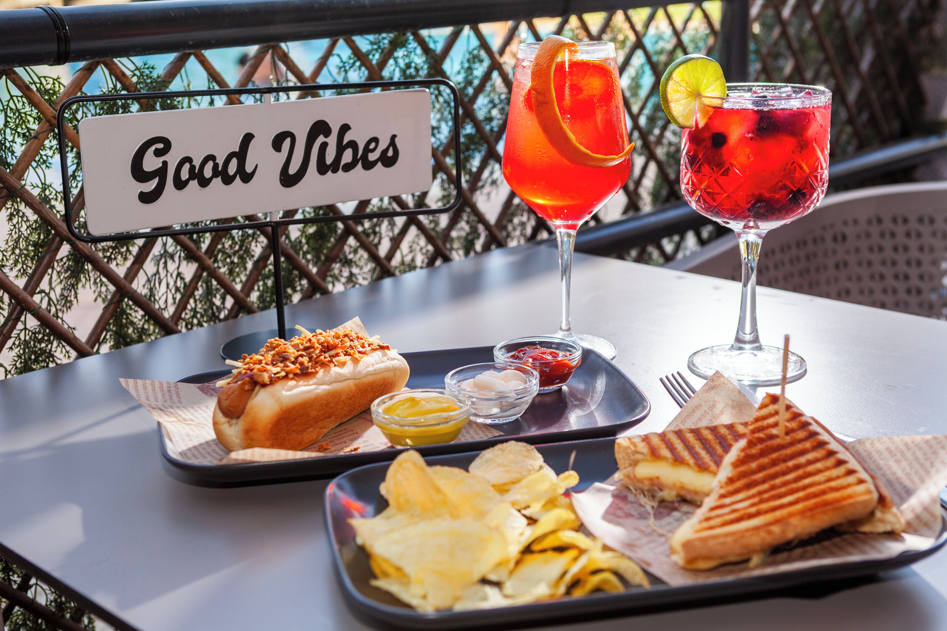 Una mesa con un cartel que dice 'Good Vibes', un sándwich, papas fritas y dos bebidas en un entorno exterior soleado.