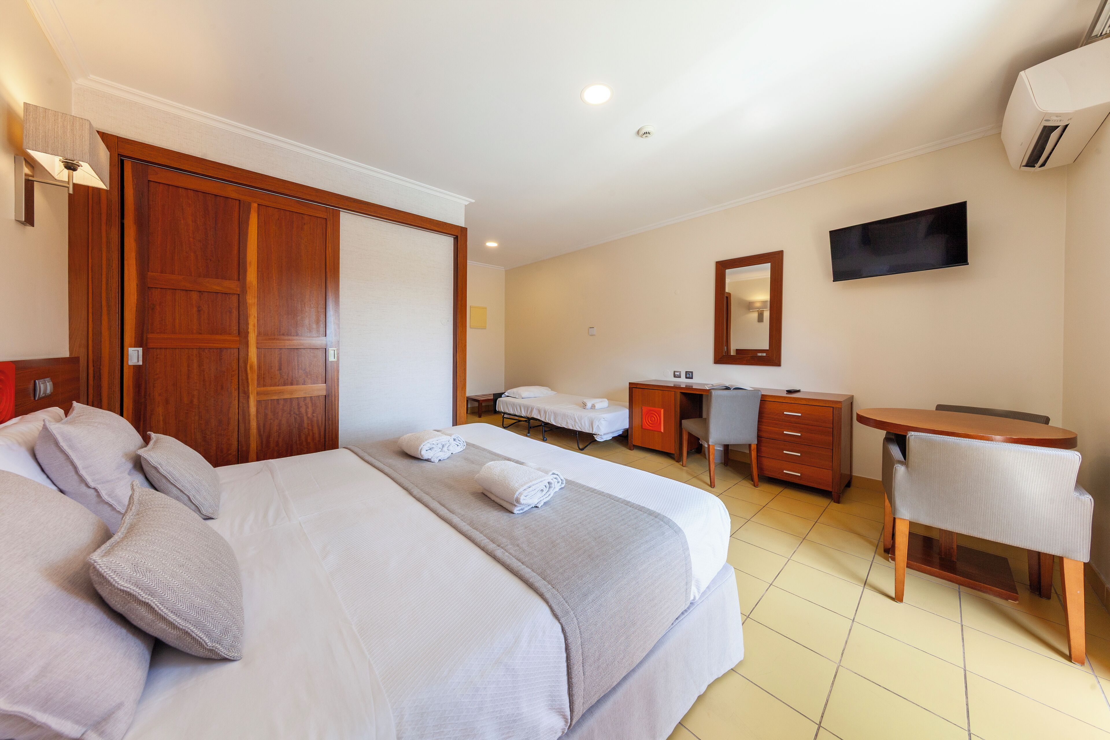 A hotel room with a large bed, a desk, a chair, a TV, and a wardrobe.