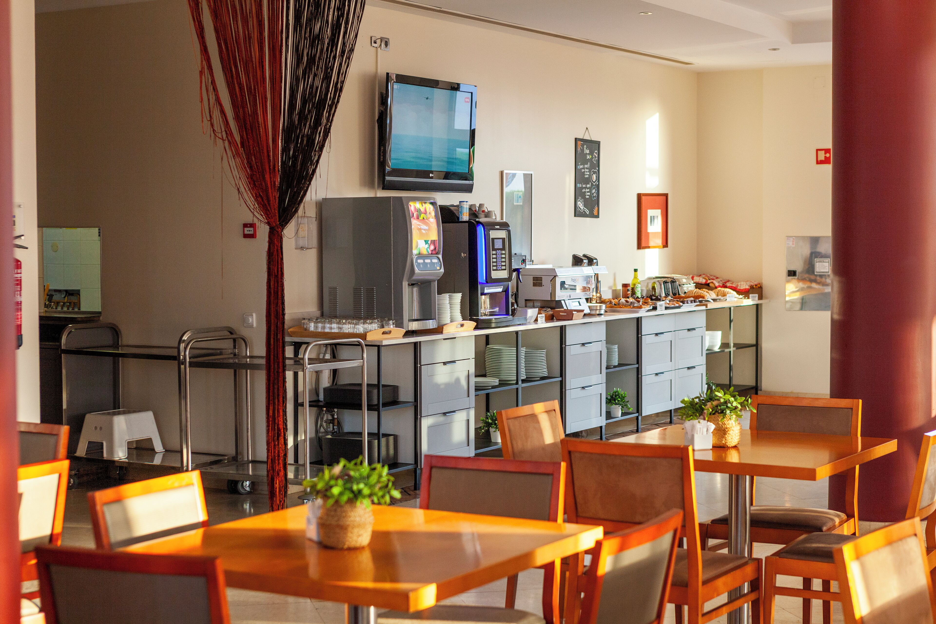 Un comedor de hotel con mesas y sillas, una zona de cocina con electrodomésticos, y un televisor montado en la pared.