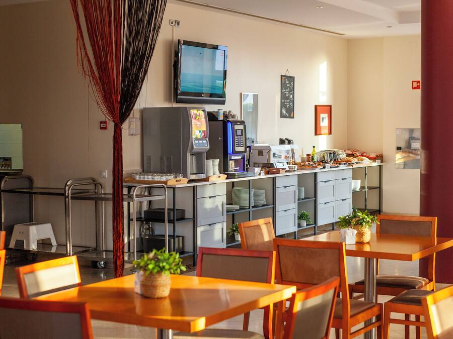 A hotel dining area with tables and chairs, a kitchen area with appliances, and a television mounted on the wall.