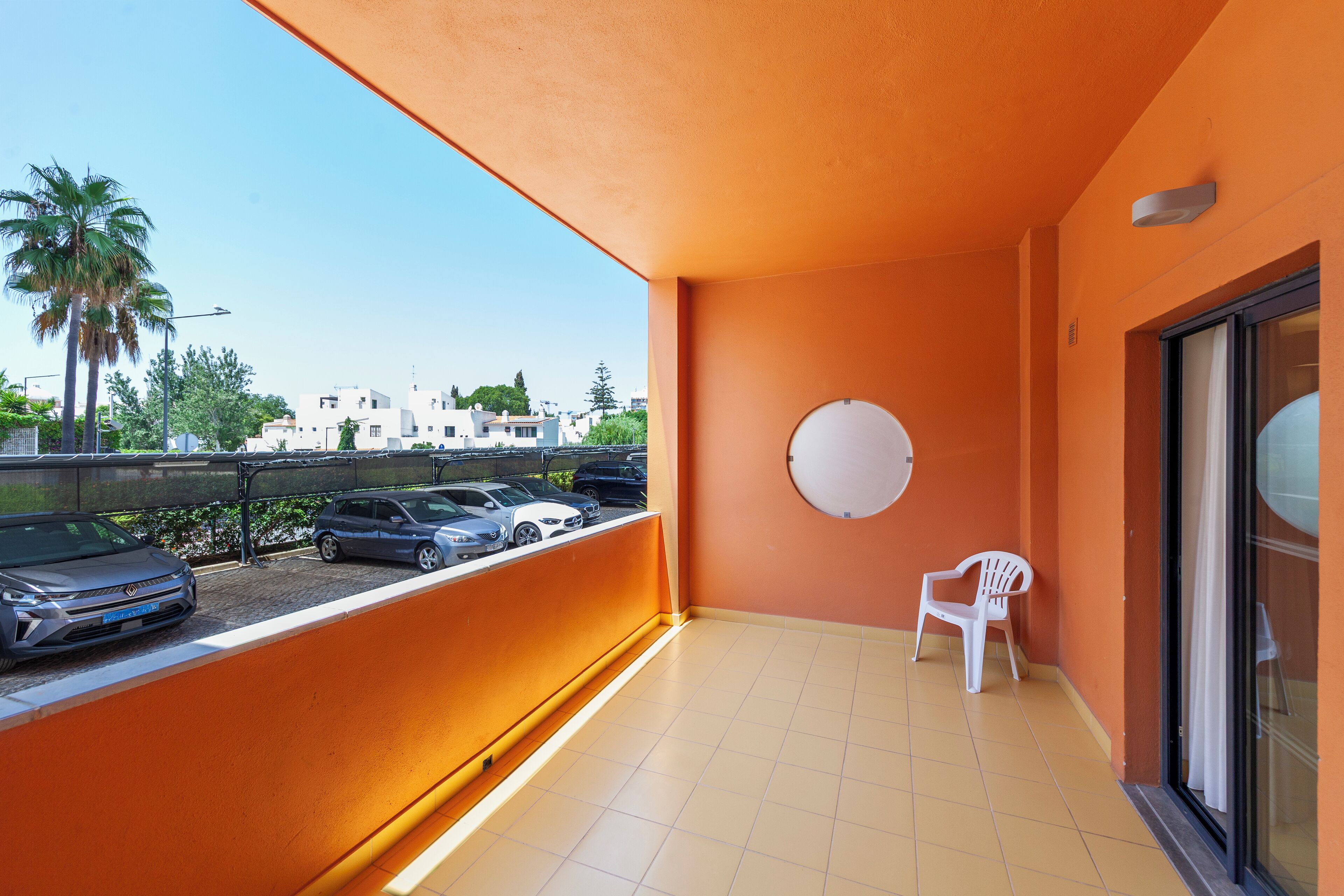 A balcony with orange walls, a white chair, and a view of parked cars and buildings.