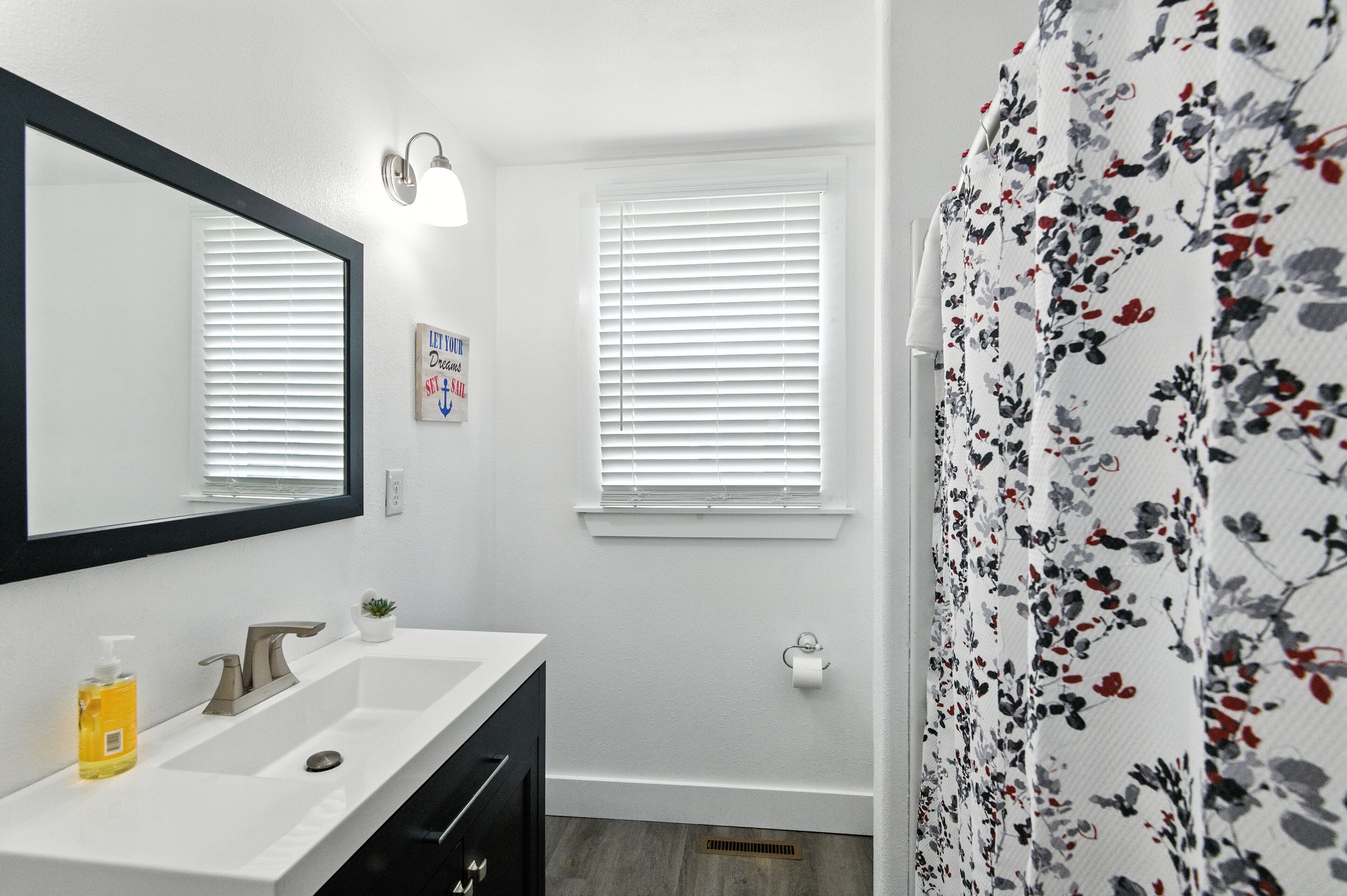 Een badkamer met een wit wastafeltje, een spiegel en een raam met jaloezieën.